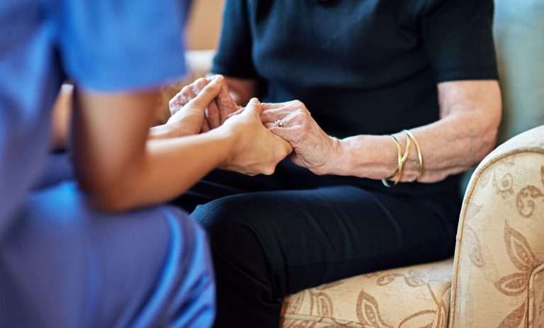A caregiver in a blue uniform gently holds the hands of an elderly person, seated on a patterned chair in an assisted living facility.