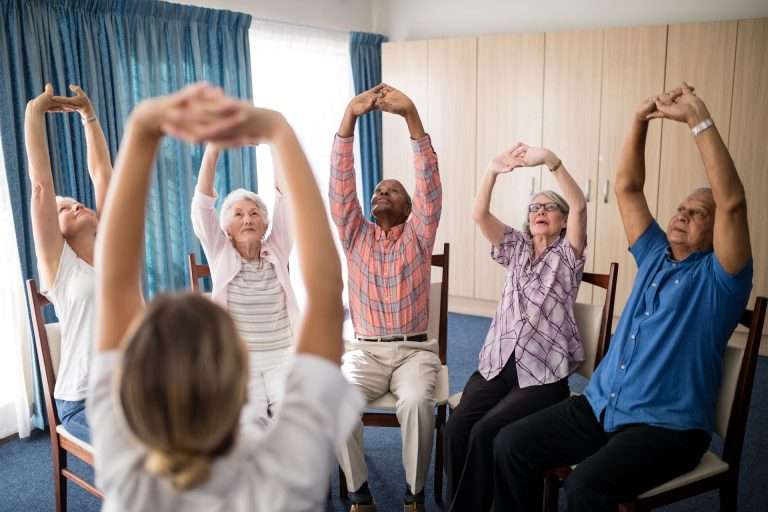 In a sunlit room, a group of elderly individuals in an assisted living facility sit on chairs as an instructor guides them to stretch their arms overhead.