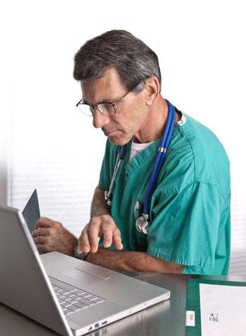 During a site visit, a doctor in green scrubs and a stethoscope efficiently types on a laptop while holding an X-ray, ensuring thorough EHR due diligence.